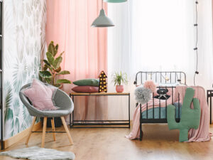 Pink pillow on grey armchair near bed in girl's bedroom interior with green cactus and lights on table